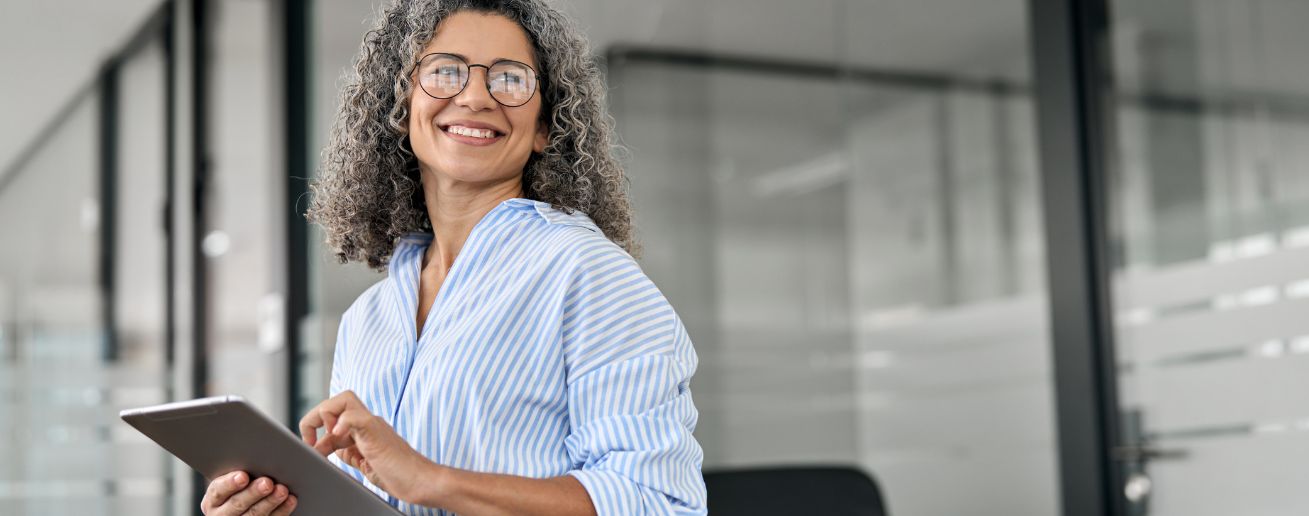 A curly-haired woman wearing glasses smiling warmly at the camera holding a tablet in her hands.