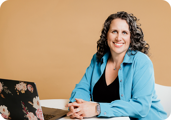 Coach Shannon sits with her hands folded over a book, smiling in a blue button-down shirt and black v-neck tee. 